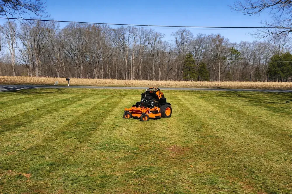 Orange riding mower cutting grass in a large green field with trees in the background