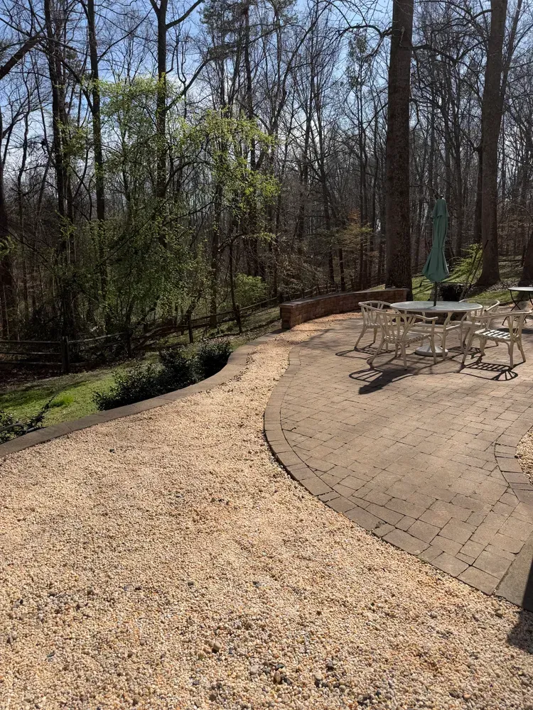 Curved gravel path beside a brick patio with metal table and chairs in a wooded backyard