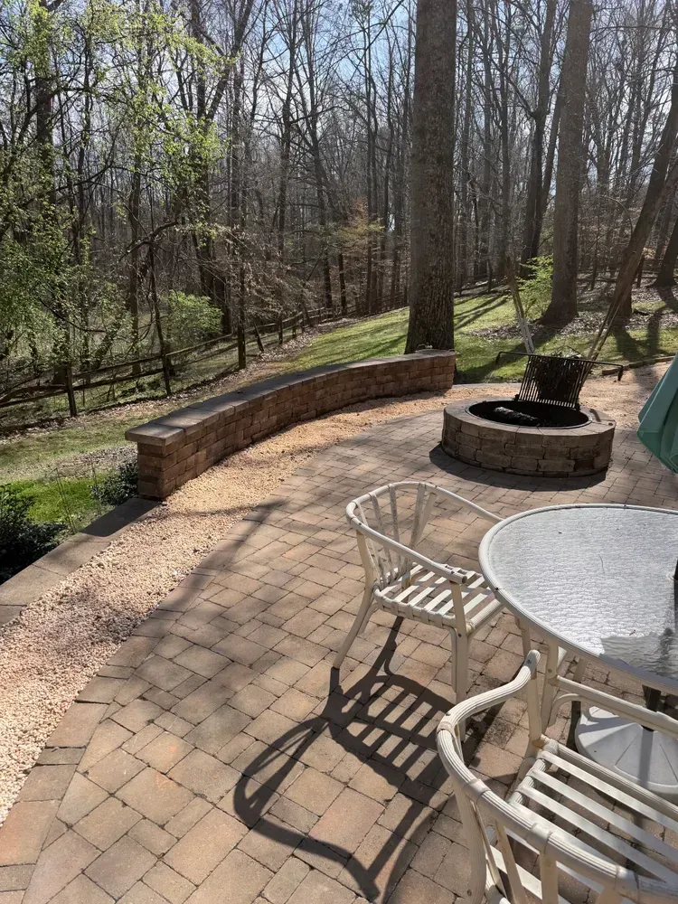 Brick patio with white table and chairs, stone fire pit, and curved retaining wall beside a wooded yard