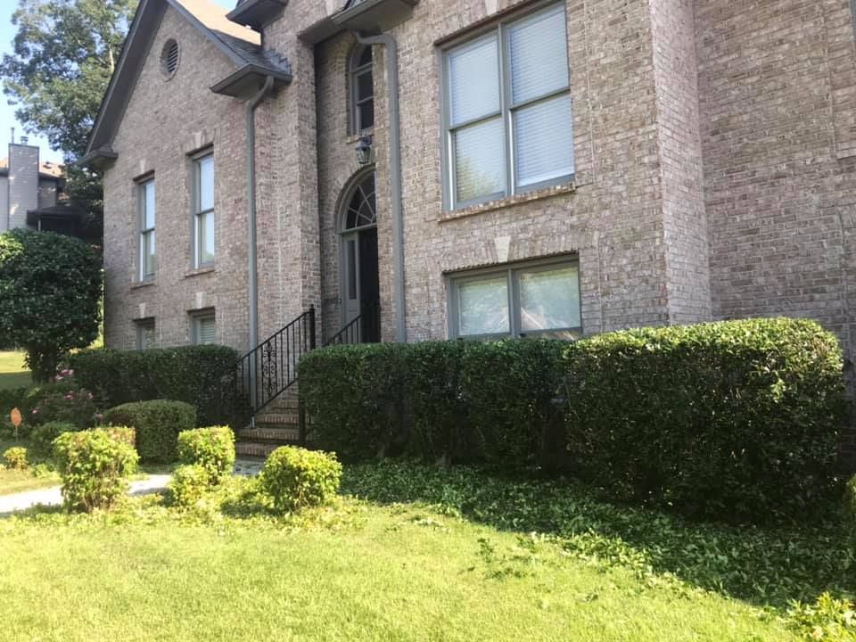 Brick apartment building with black iron entrance, windows, and trimmed hedges beside a green lawn.