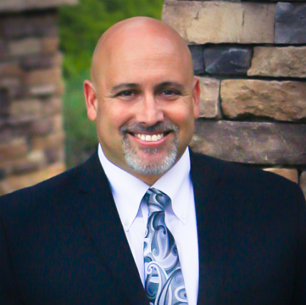 A bald man in a suit and tie is smiling in front of a stone wall.