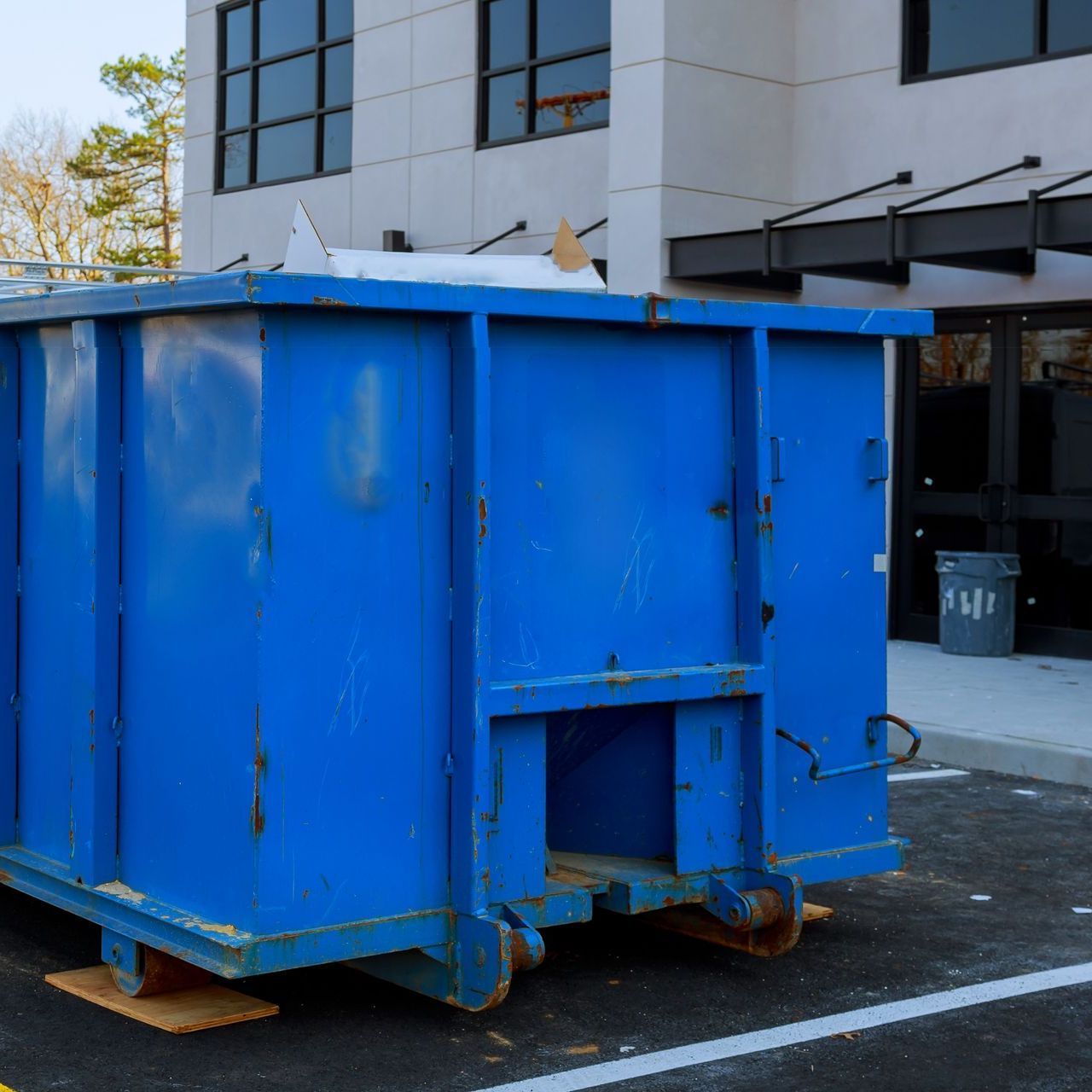 A blue dumpster is parked in a parking lot in front of a building.