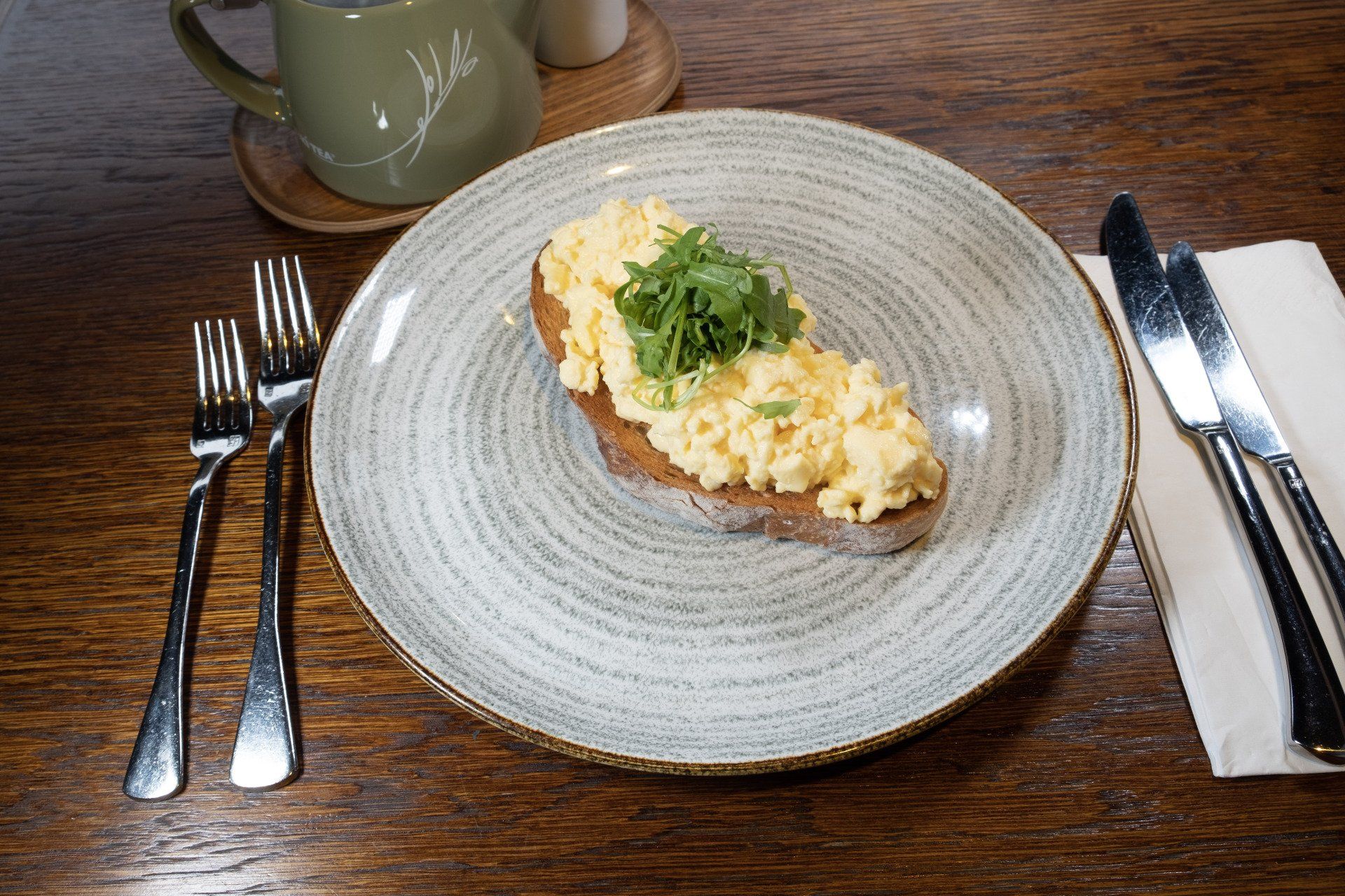 Scrambled eggs on toast, garnished with microgreens, on a plate with silverware, next to a mug on a wood table.