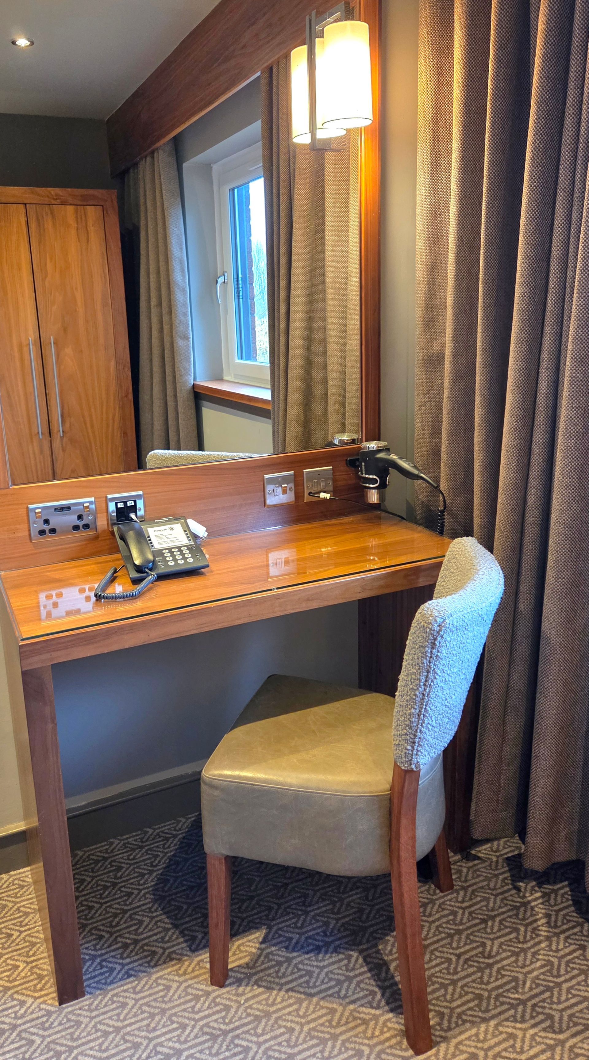Hotel room desk with mirror, kettle, phone, and chair next to a window and curtain.