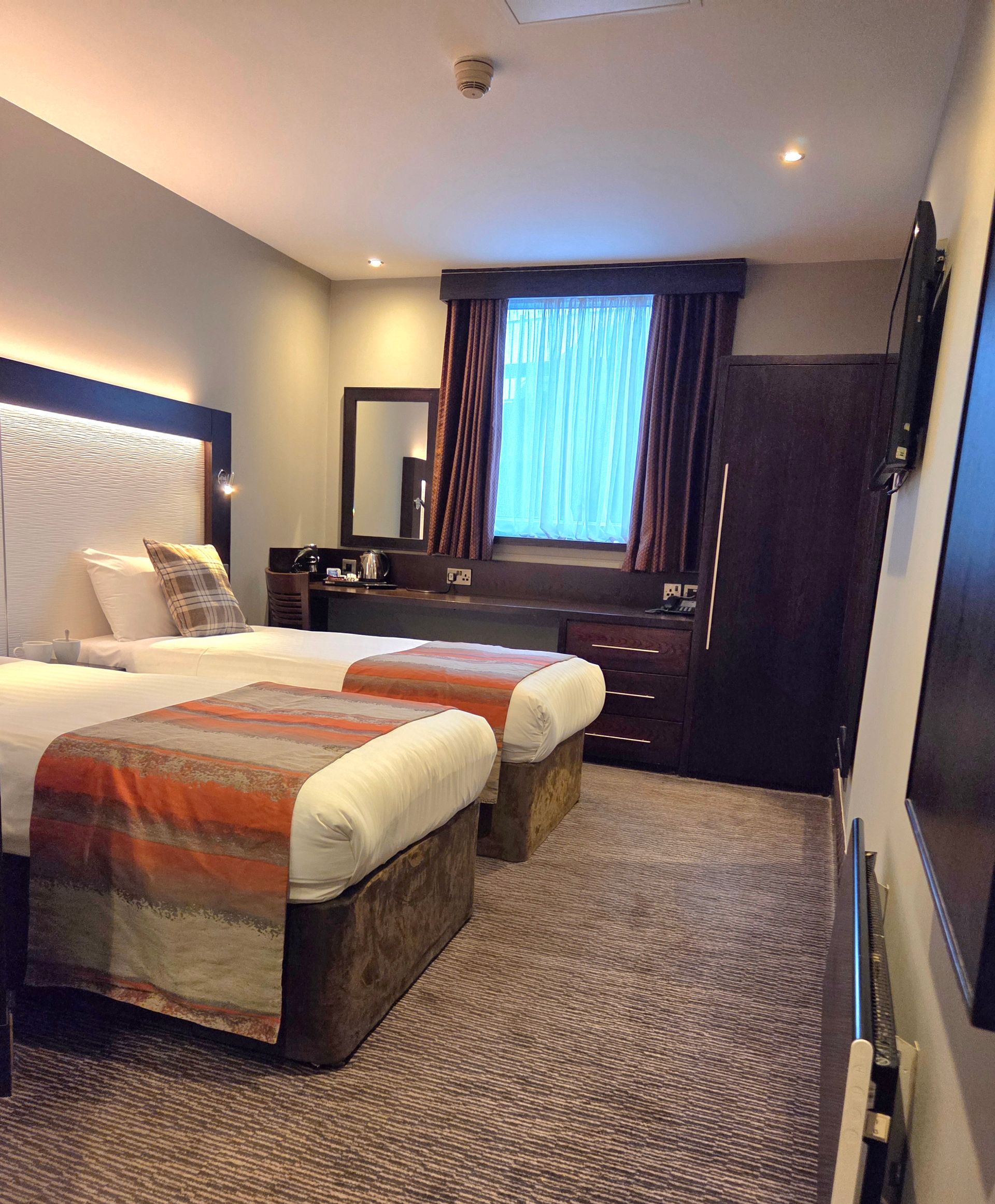 A hotel room with two twin beds, desk, wardrobe, and window with plaid accents.