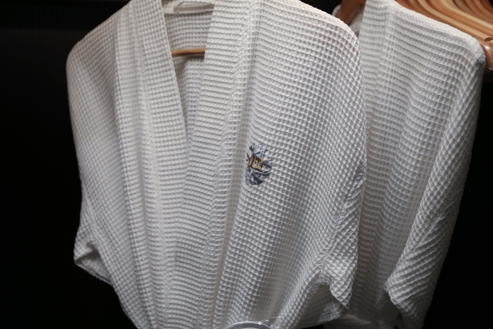 Two white waffle-weave bathrobes hanging on wooden hangers in a dark closet. One has a small embroidered emblem.