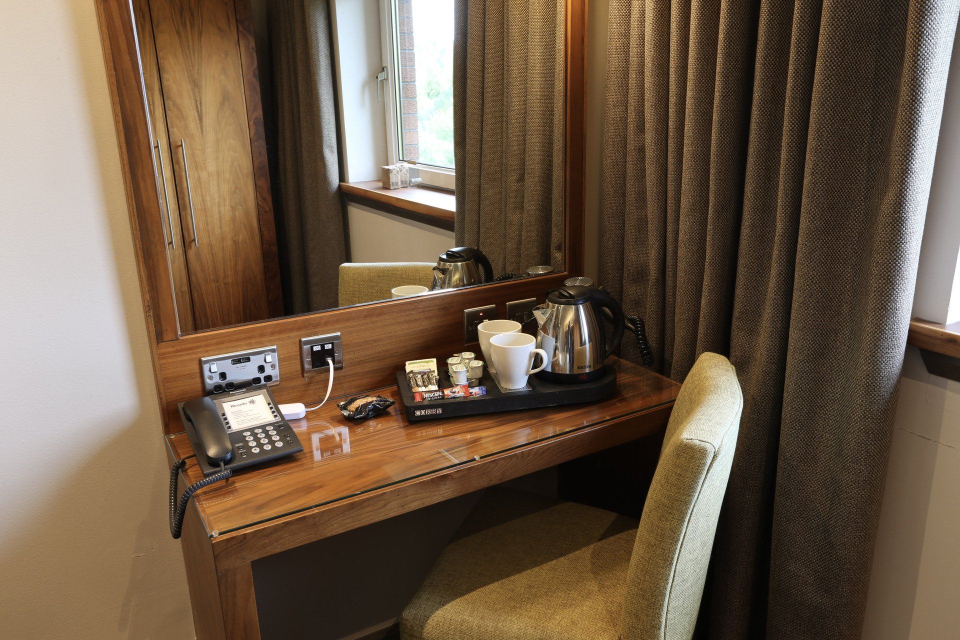 Hotel room desk with mirror, kettle, phone, and chair next to a window and curtain.