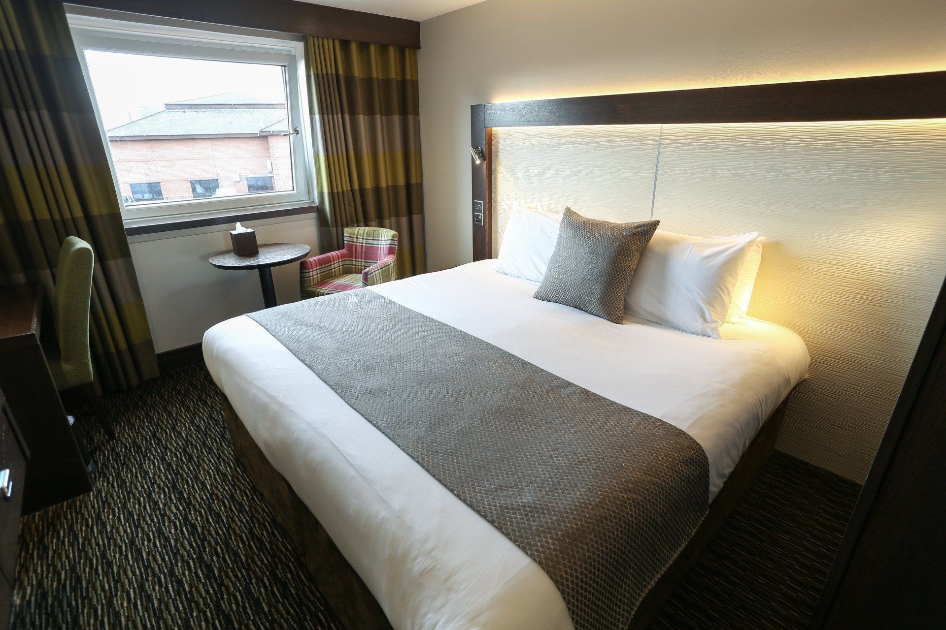 Hotel room with a queen bed, window, chair, and dark patterned carpet.
