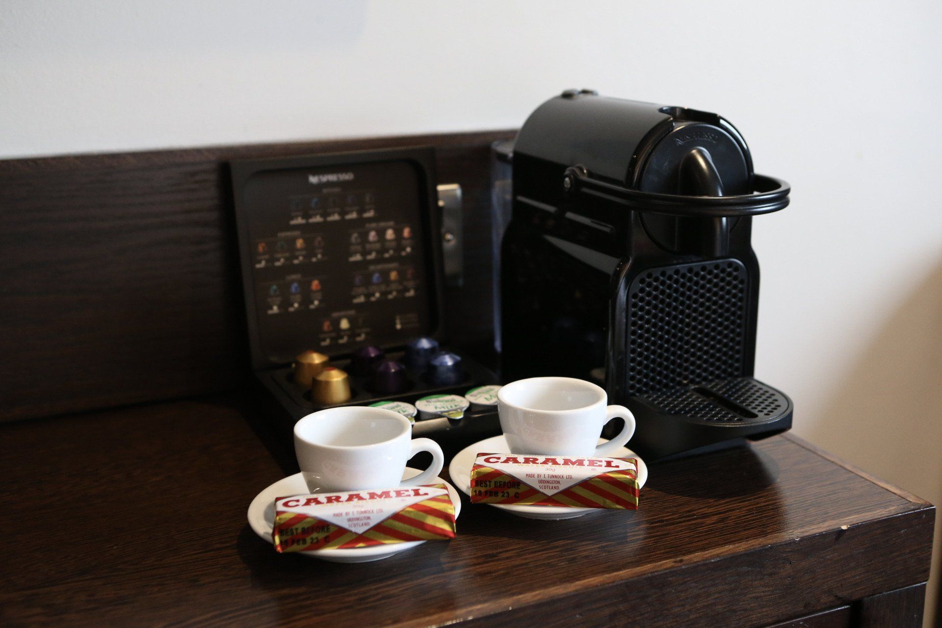 A coffee station with a black Nespresso machine, cups, pods, and caramel biscuits on a dark wooden surface.