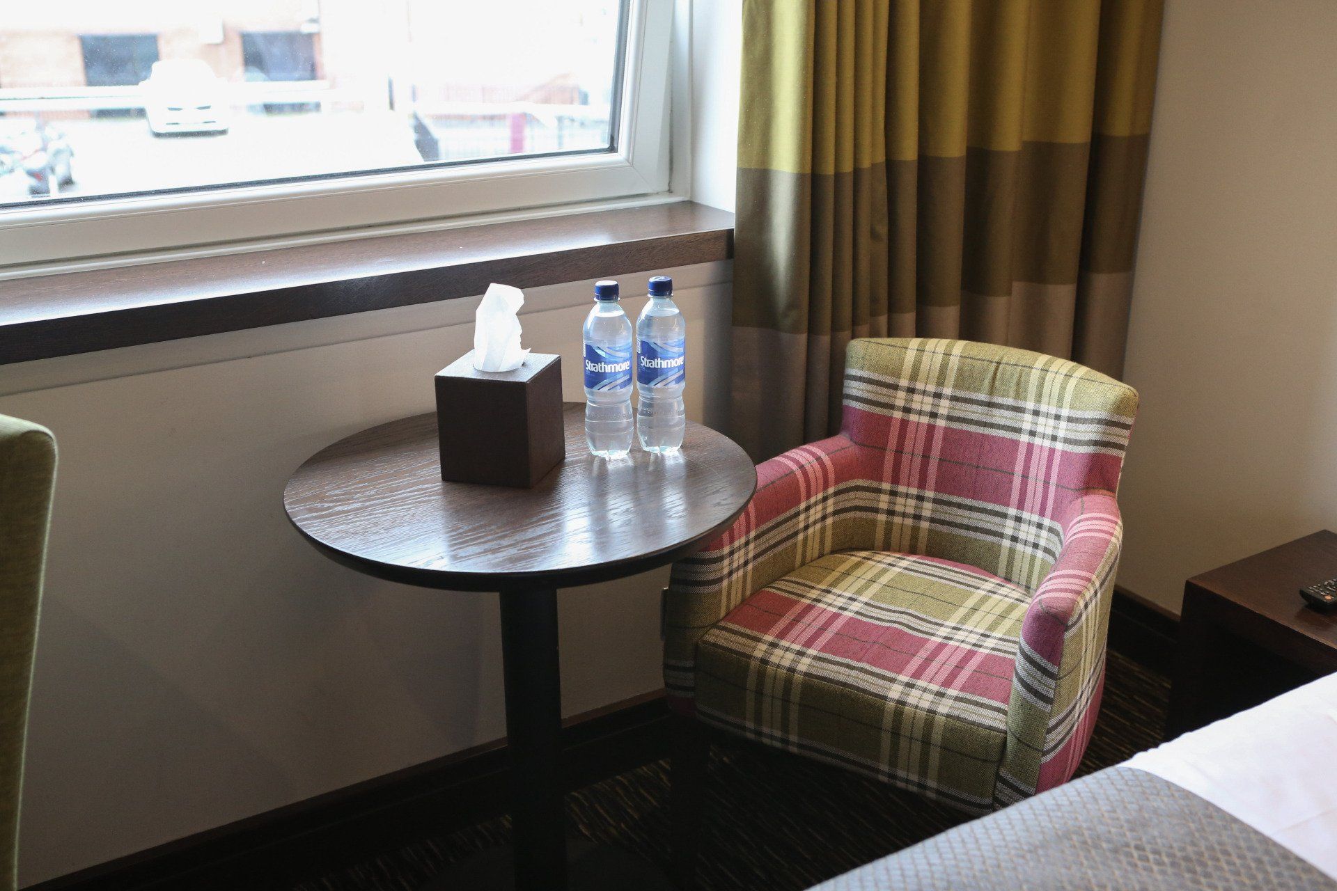 A hotel room with a plaid armchair, small table with water bottles and tissues, and a window.