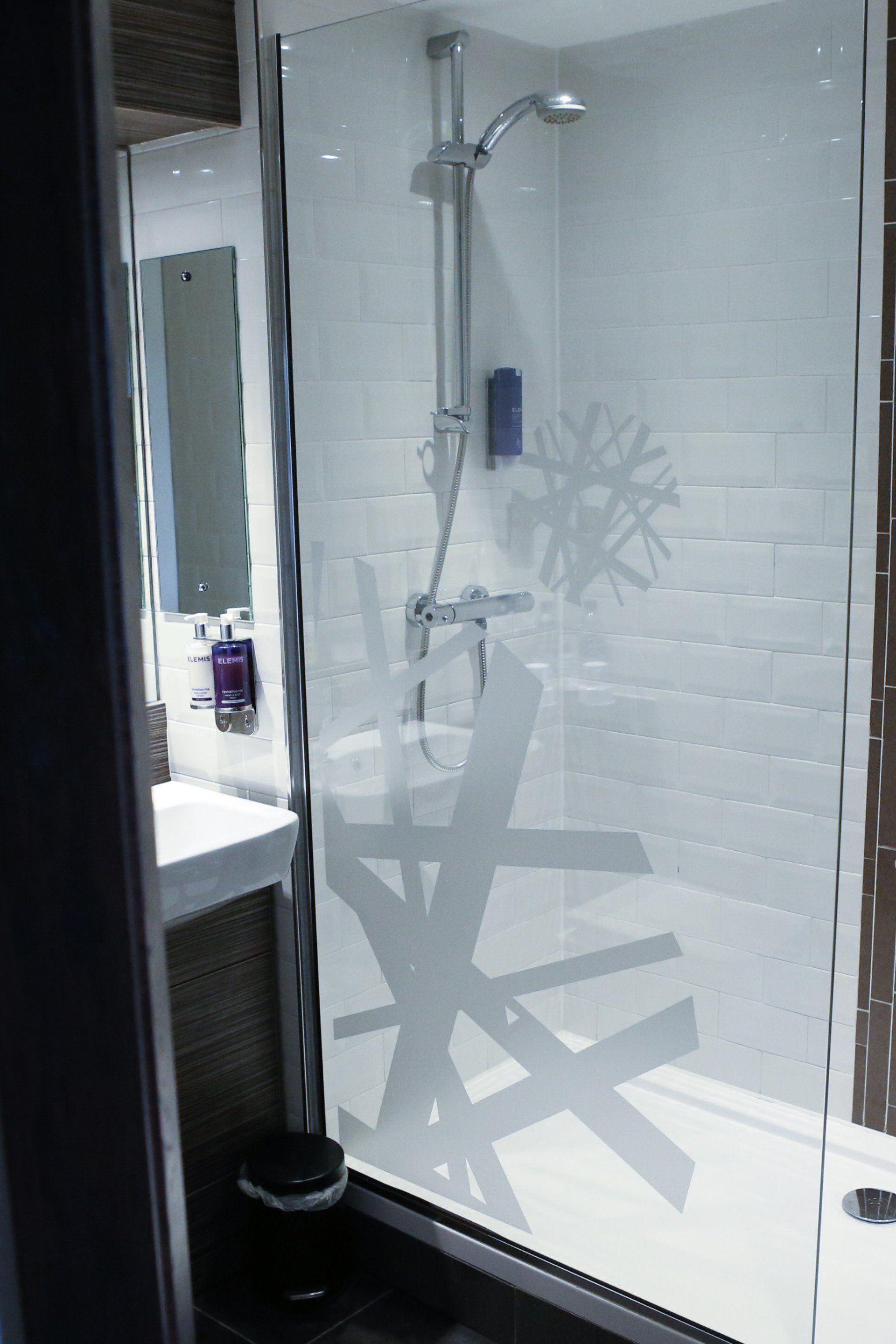 Shower with glass door, white tiled walls, and shower head. Bathroom has a sink and trash can.