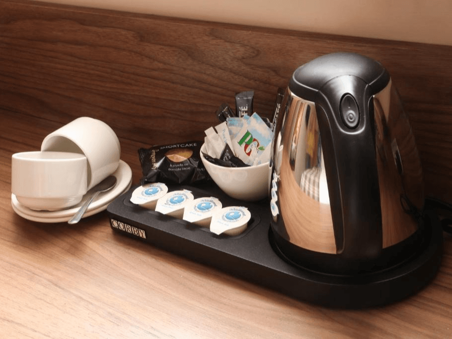 Hotel room coffee station with kettle, cups, tea, and sweeteners on a wooden surface.