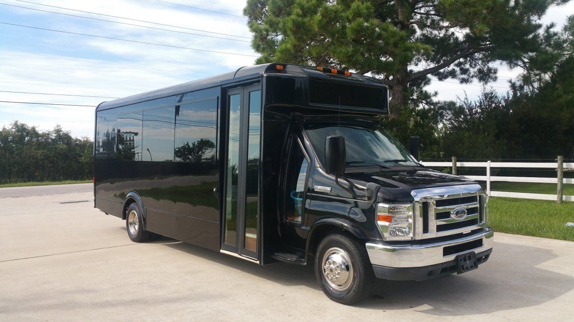 Black party bus parked on a paved area, in front of greenery and a white fence.