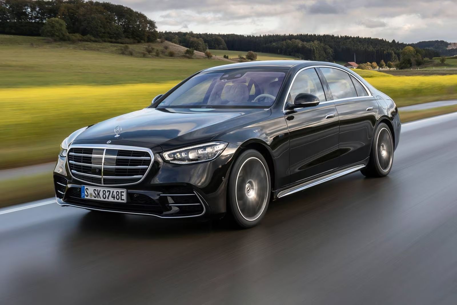 Black Mercedes-Benz S-Class sedan driving on a road with a green field and hills in the background.