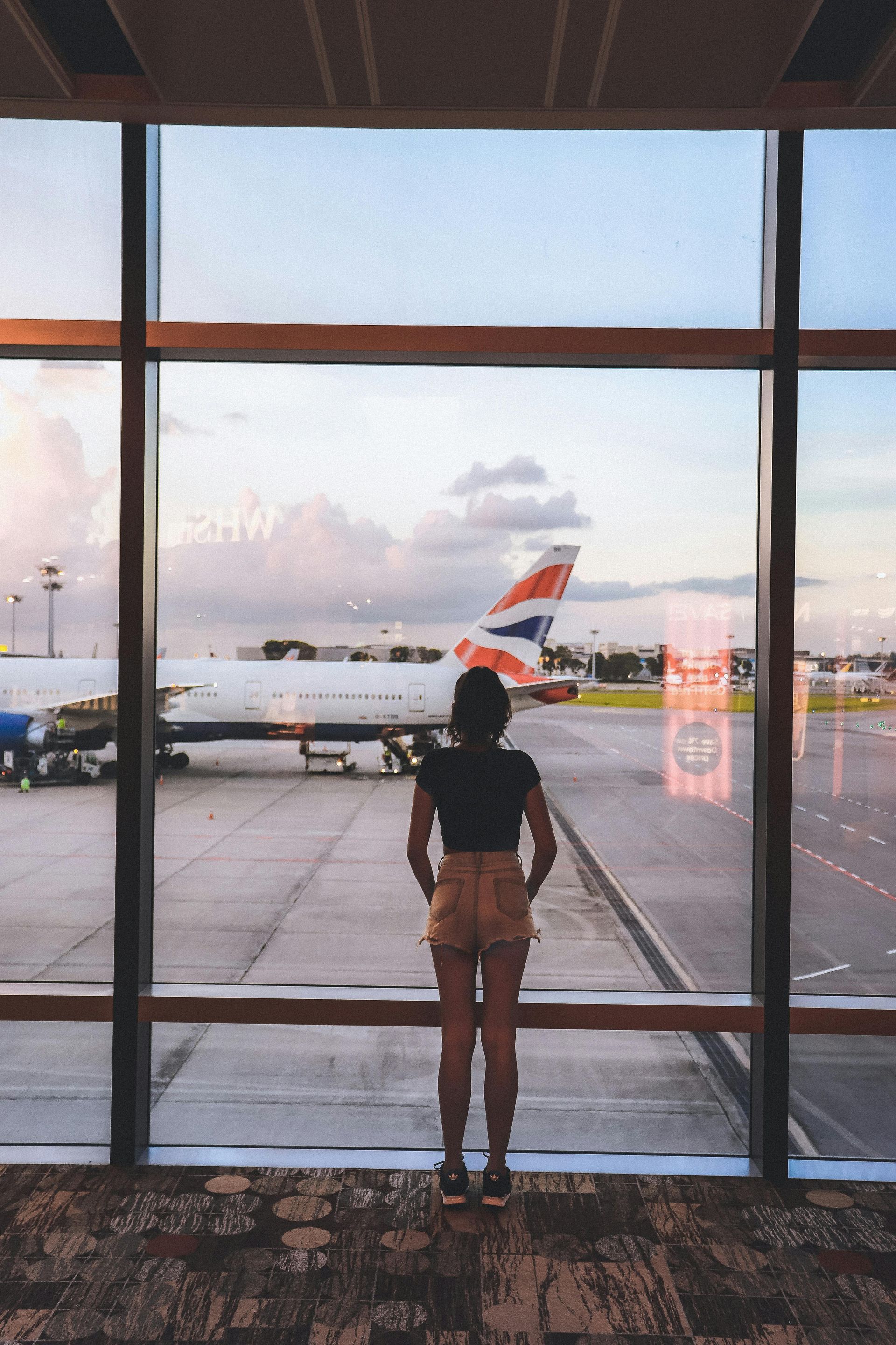 Woman at airport window watches plane. Sky is pink and cloudy.