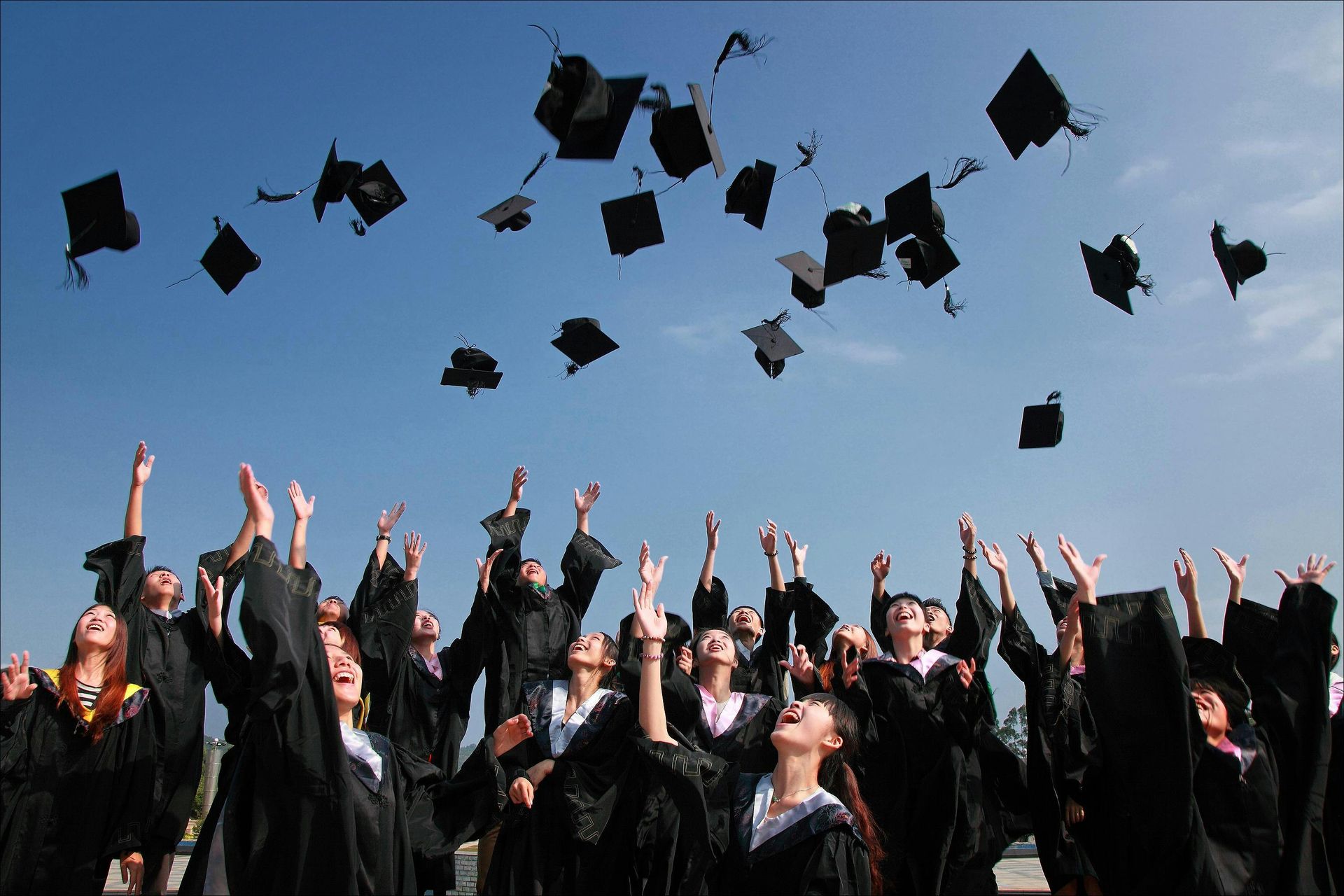 Graduates in black gowns tossing mortarboards in the air against a bright blue sky.