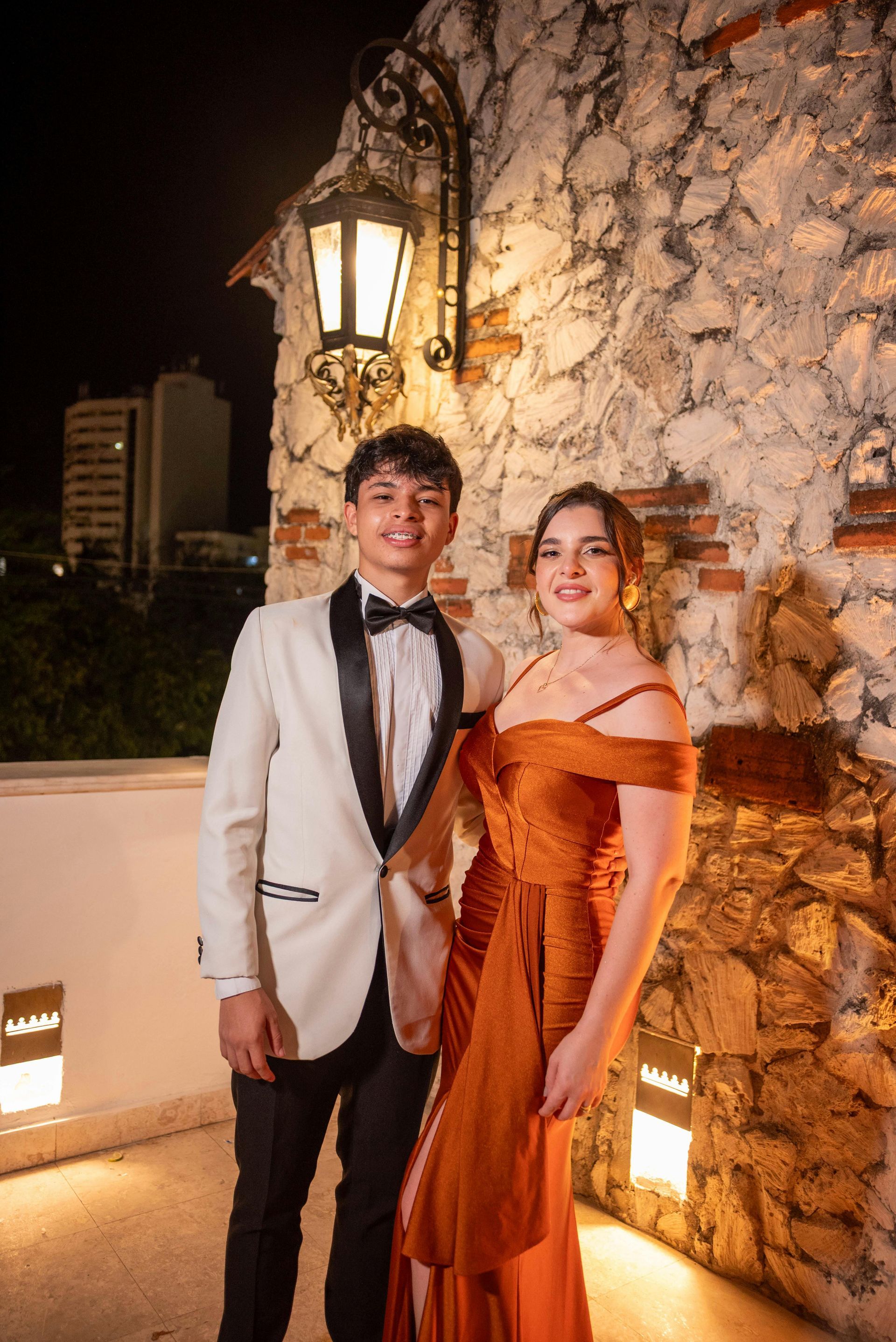 Young couple in formal wear posing outdoors, woman in orange gown, man in cream tuxedo, stone wall.