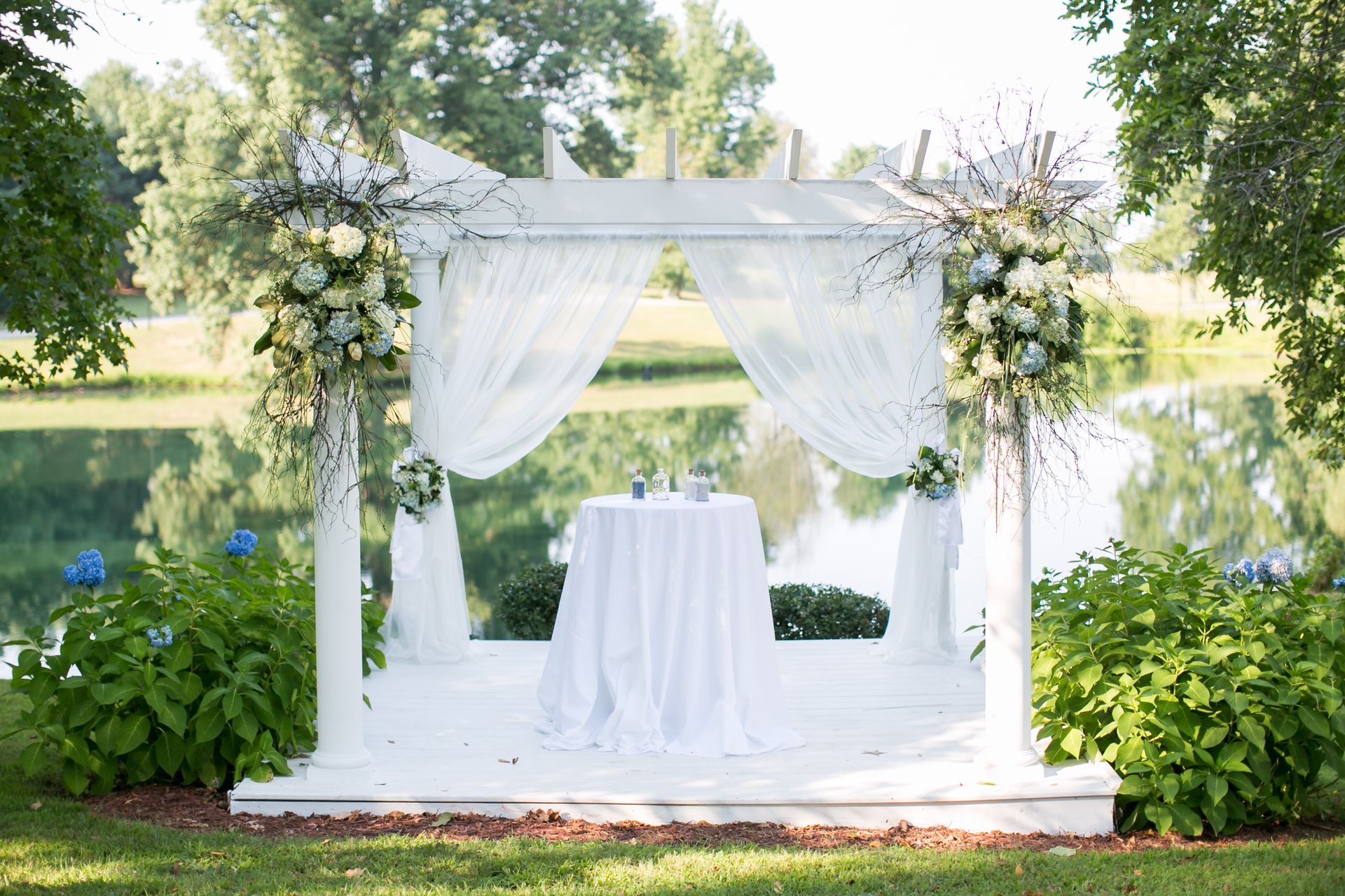 A white gazebo with flowers and a table in front of a lake.