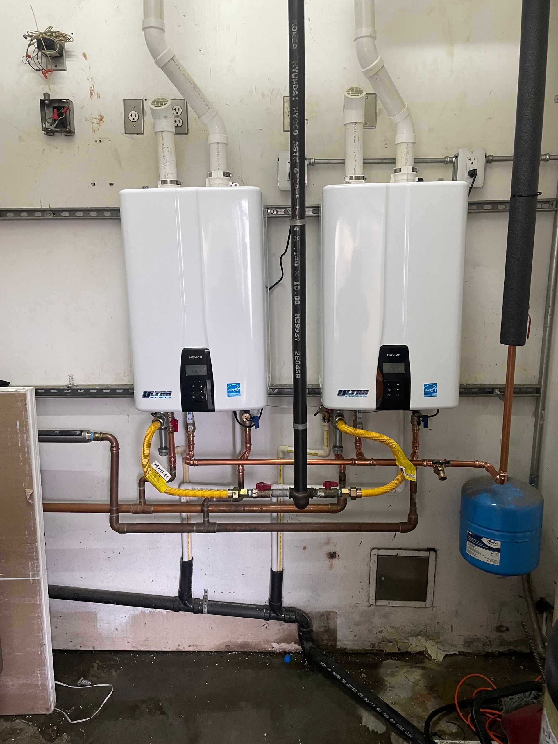 Two water heaters are connected to copper pipes in a room.