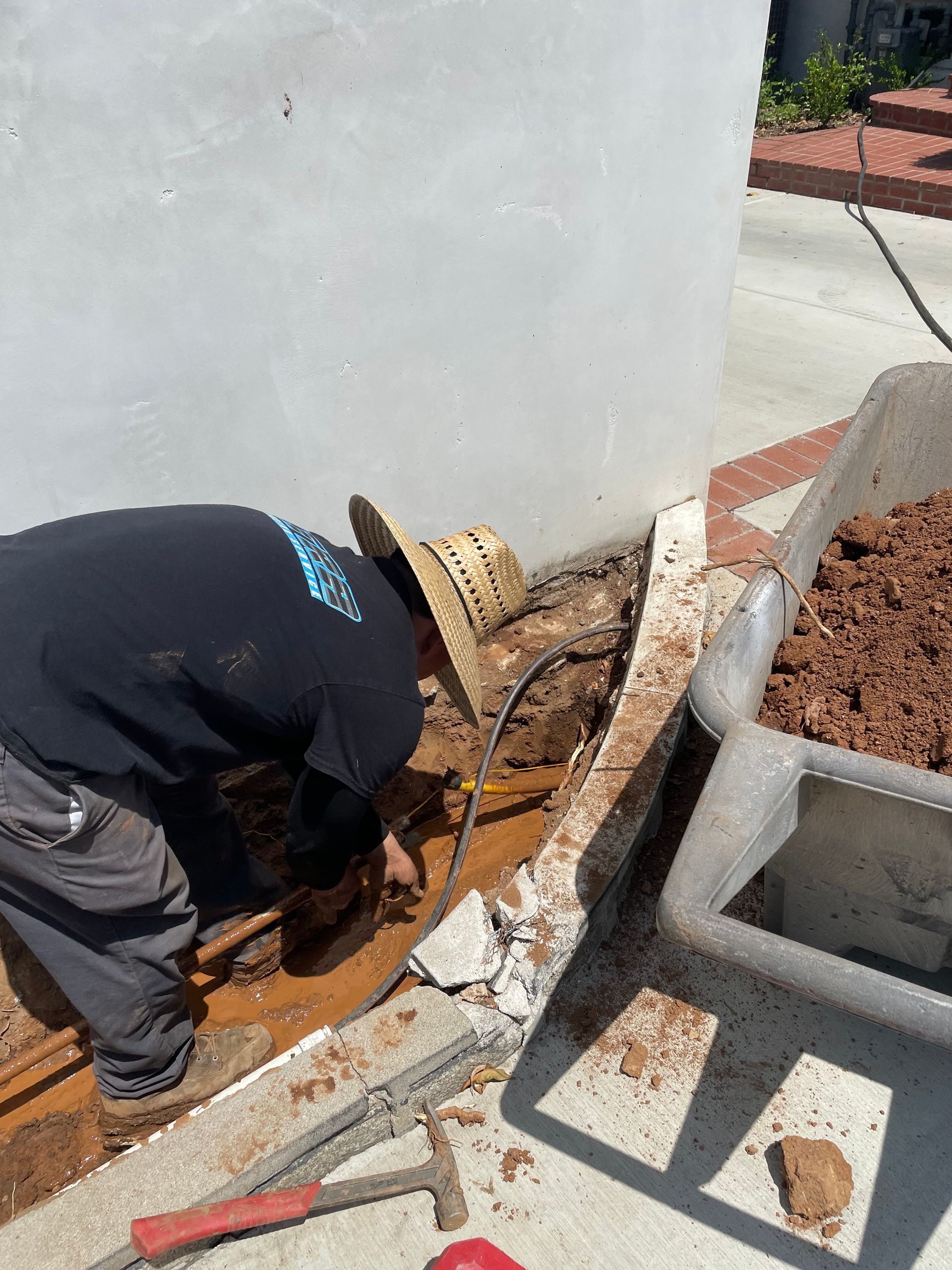 A man wearing a straw hat is working on a sidewalk next to a wheelbarrow filled with dirt.
