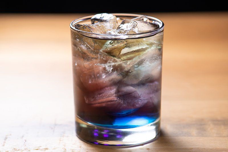 A glass of purple and blue liquid with ice on a wooden table.