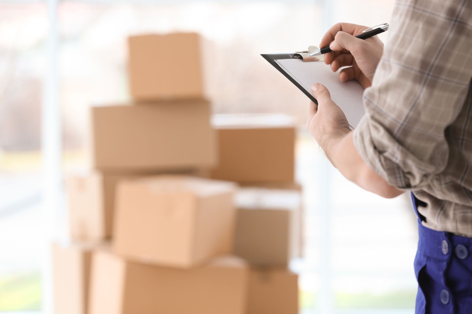 Person writing on a clipboard beside stacked cardboard boxes in a bright room