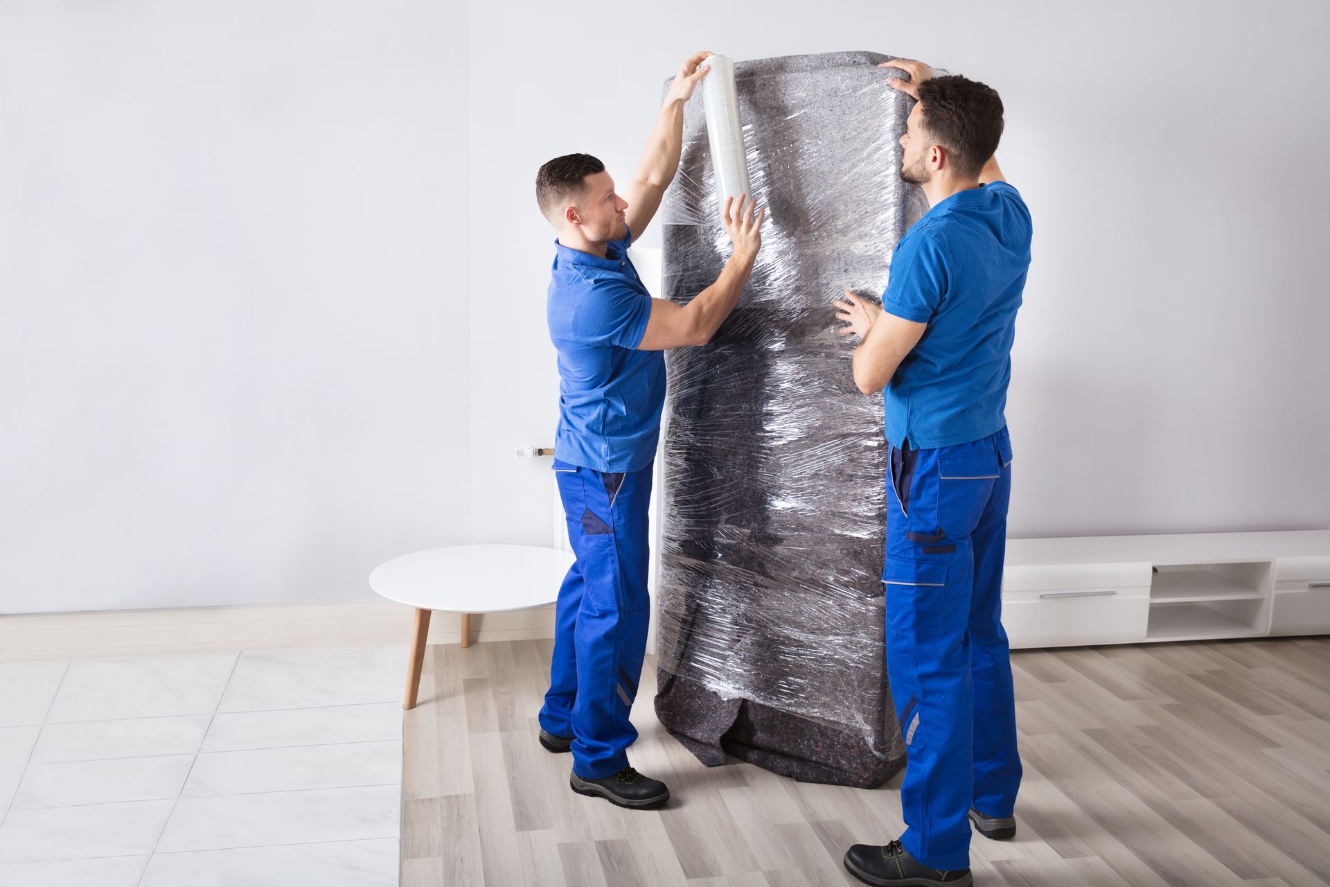 Two movers in blue uniforms wrapping a tall object in protective plastic indoors