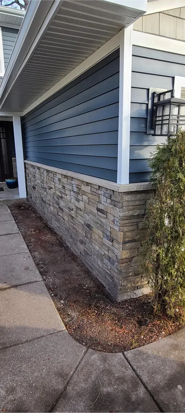 A house with a blue siding and a stone wall next to a sidewalk.