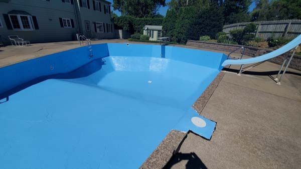 Empty blue swimming pool on a sunny day; slide, house, and yard in the background.