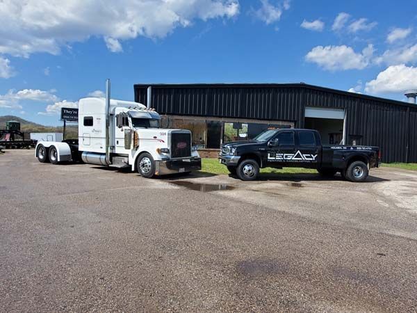 White semi-truck and black pickup truck parked in front of a dark building on a sunny day.