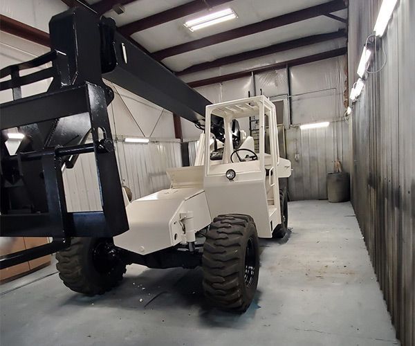 Off-white forklift in a workshop, black forks, large tires, with a black overhead arm.
