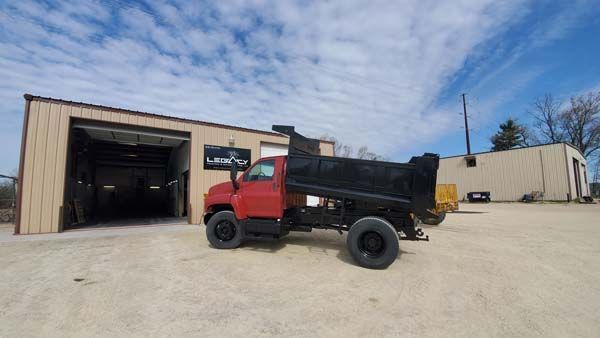 Red dump truck parked in front of a tan industrial building under a partly cloudy blue sky.