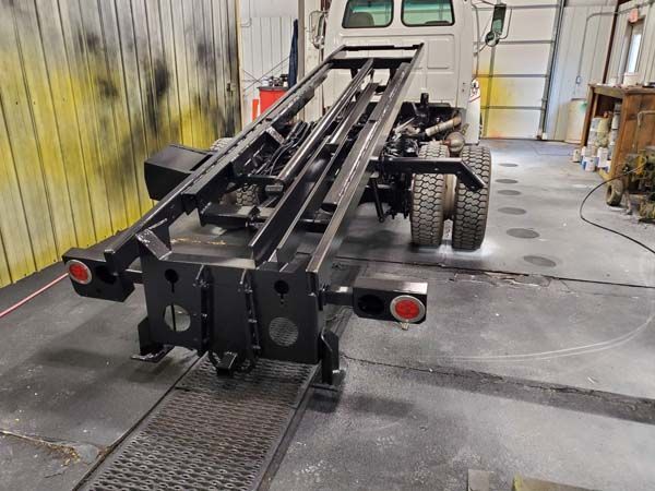 Black roll-off truck chassis on a concrete floor inside a workshop. Red taillights.