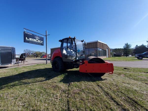 Road roller on grass, near a sign and building, under a blue sky.