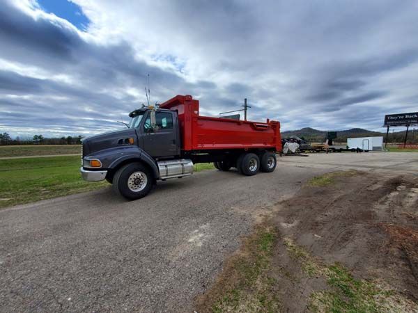 Gray dump truck with a red bed parked on a paved area on a cloudy day.