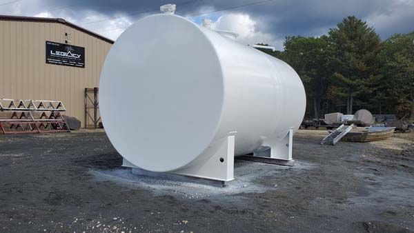White cylindrical storage tank on a metal base outdoors near a tan building and trees.