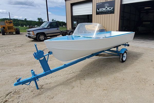 Blue and white boat on a blue trailer in front of a building, with a truck and farm equipment nearby.