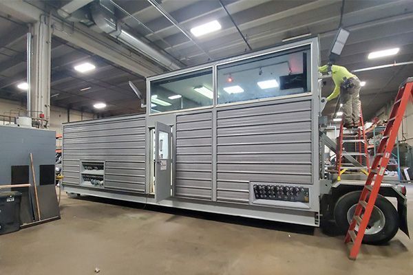 A worker on a ladder installing a window on a modern, gray prefab building inside a factory.