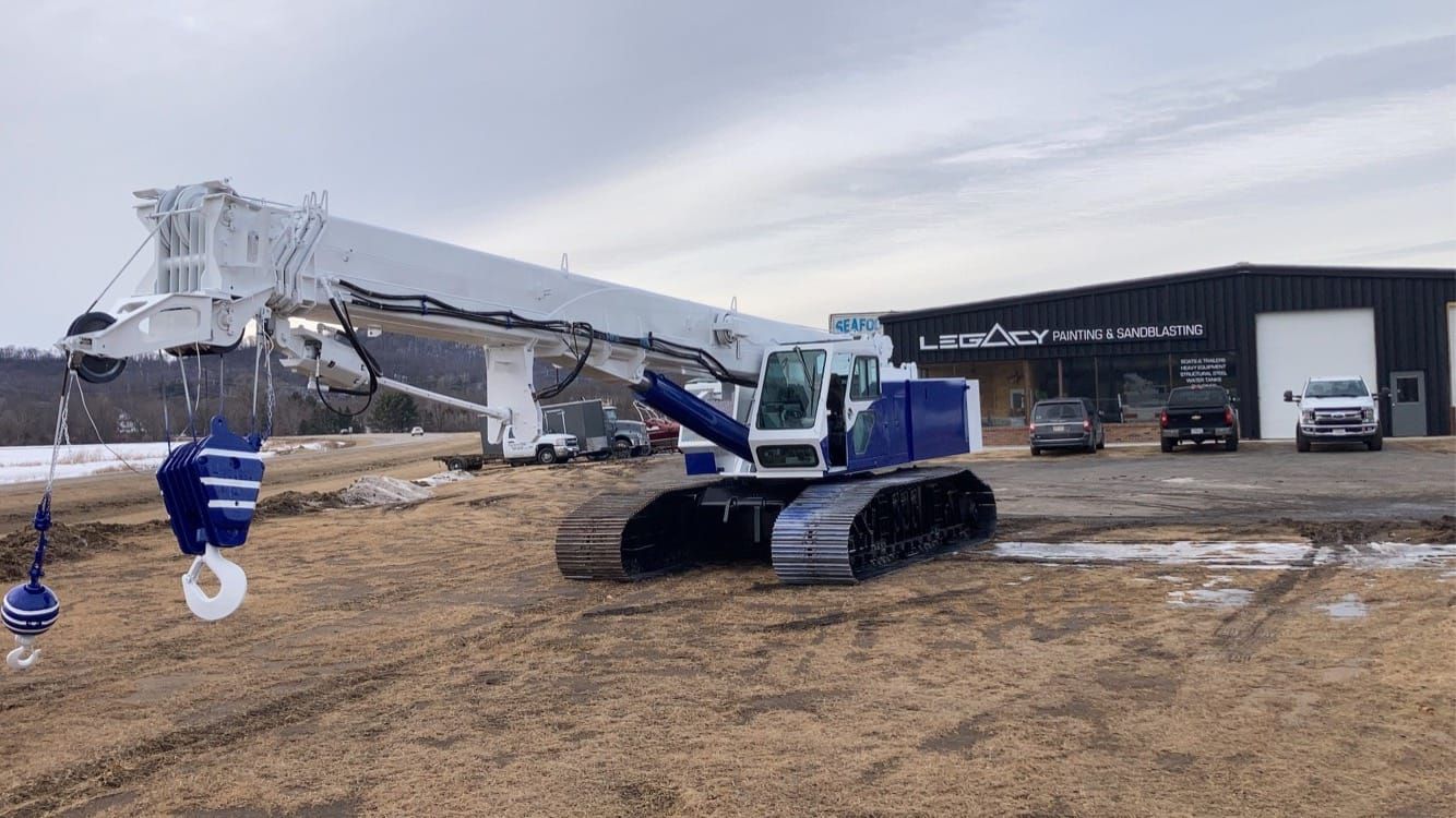 Blue and white crane on tracks in front of a building with vehicles parked outside on a cloudy day.