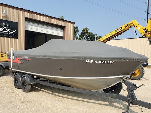 Boat covered in gray fabric on trailer; parked in front of a building with a forklift present.