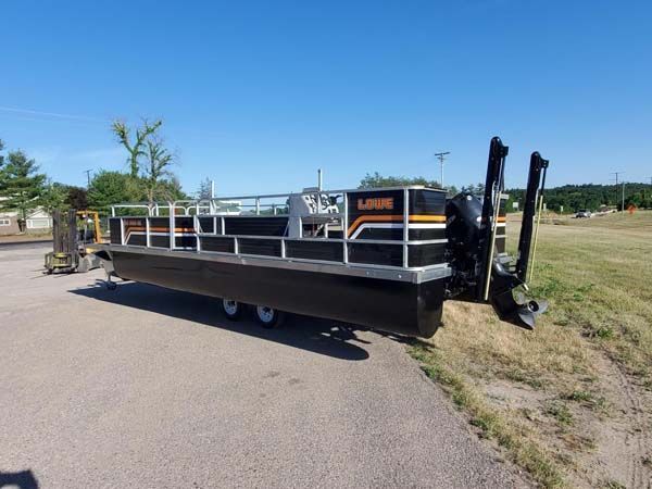 Black and orange pontoon boat on a trailer, parked on asphalt, with a blue sky.