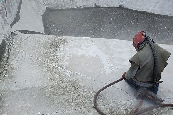 Person in safety gear sandblasting a concrete surface; dust and debris visible.