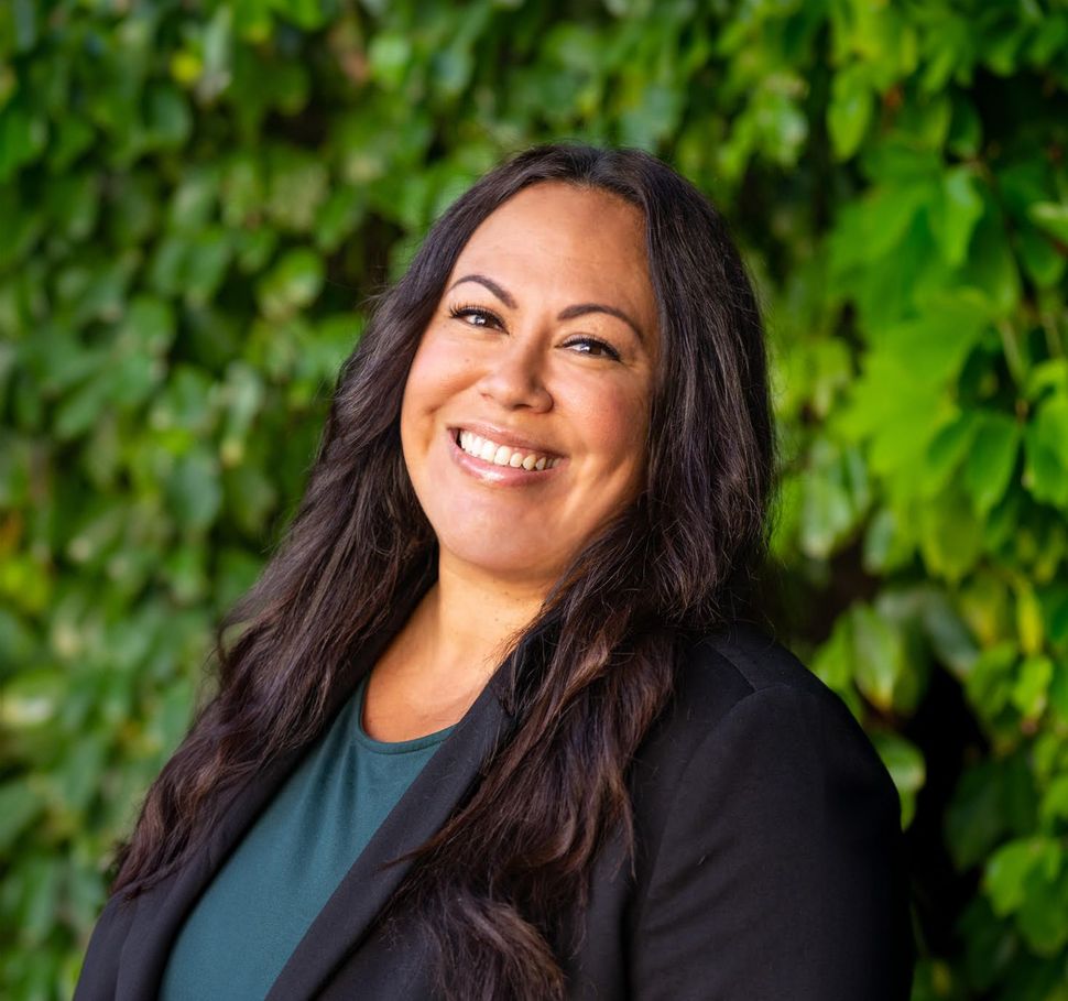 A woman in a black jacket is smiling in front of a green bush.
