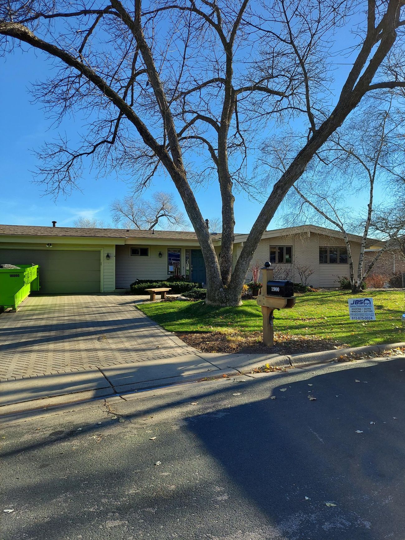 A house with a green dumpster in front of it and a tree in front of it.