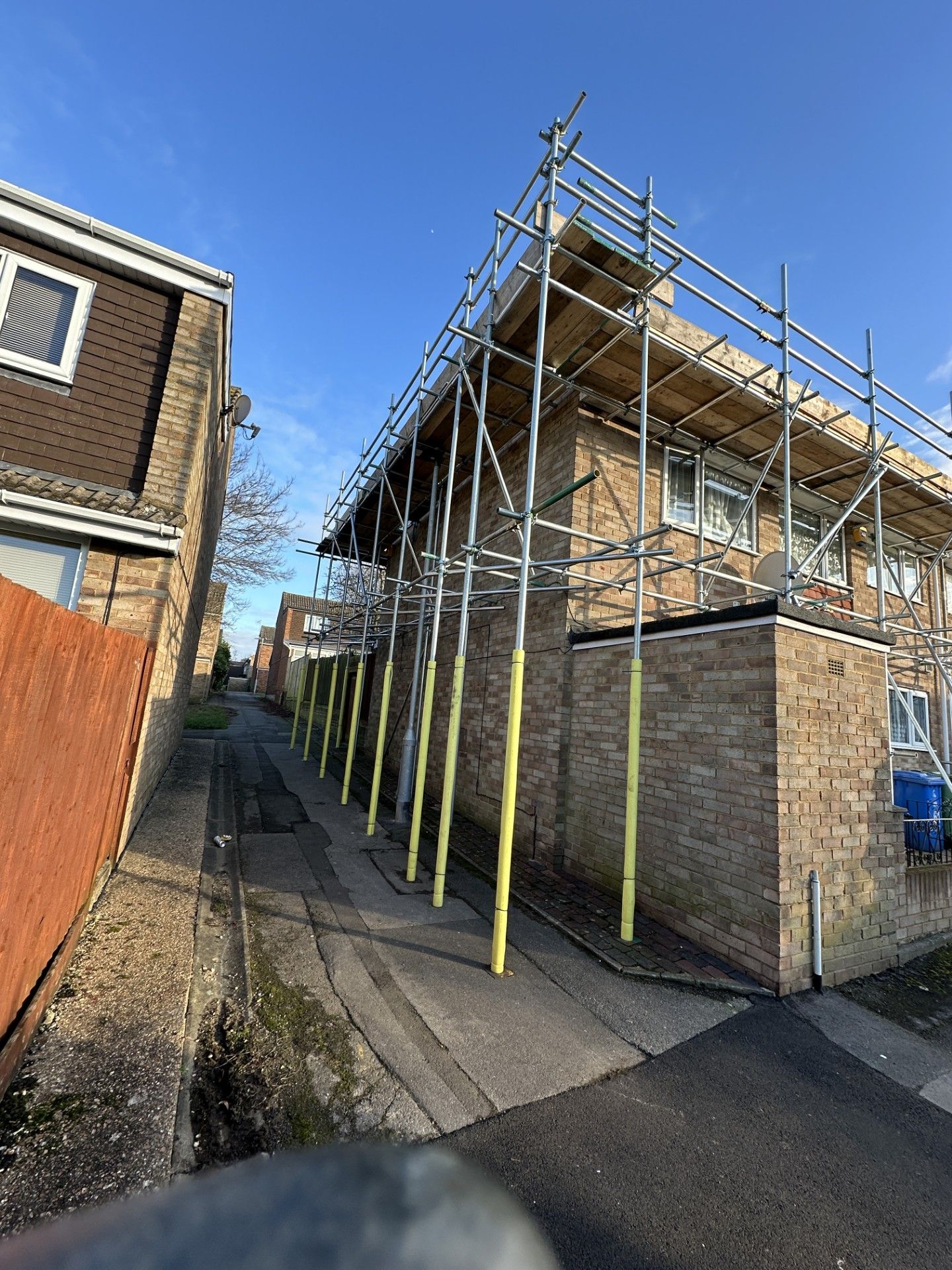 Scaffolding on a brick building's side, yellow supports, pathway to the left, sunny day.