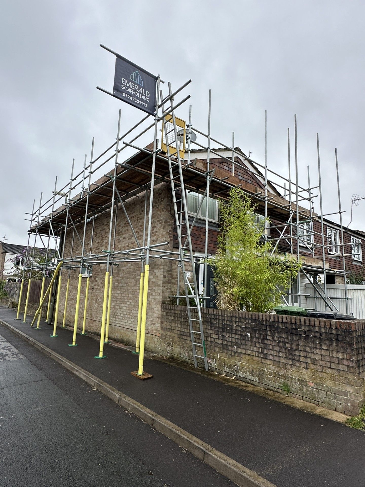 Scaffolding surrounds a brick house; cloudy day. A sign at top, street in front.