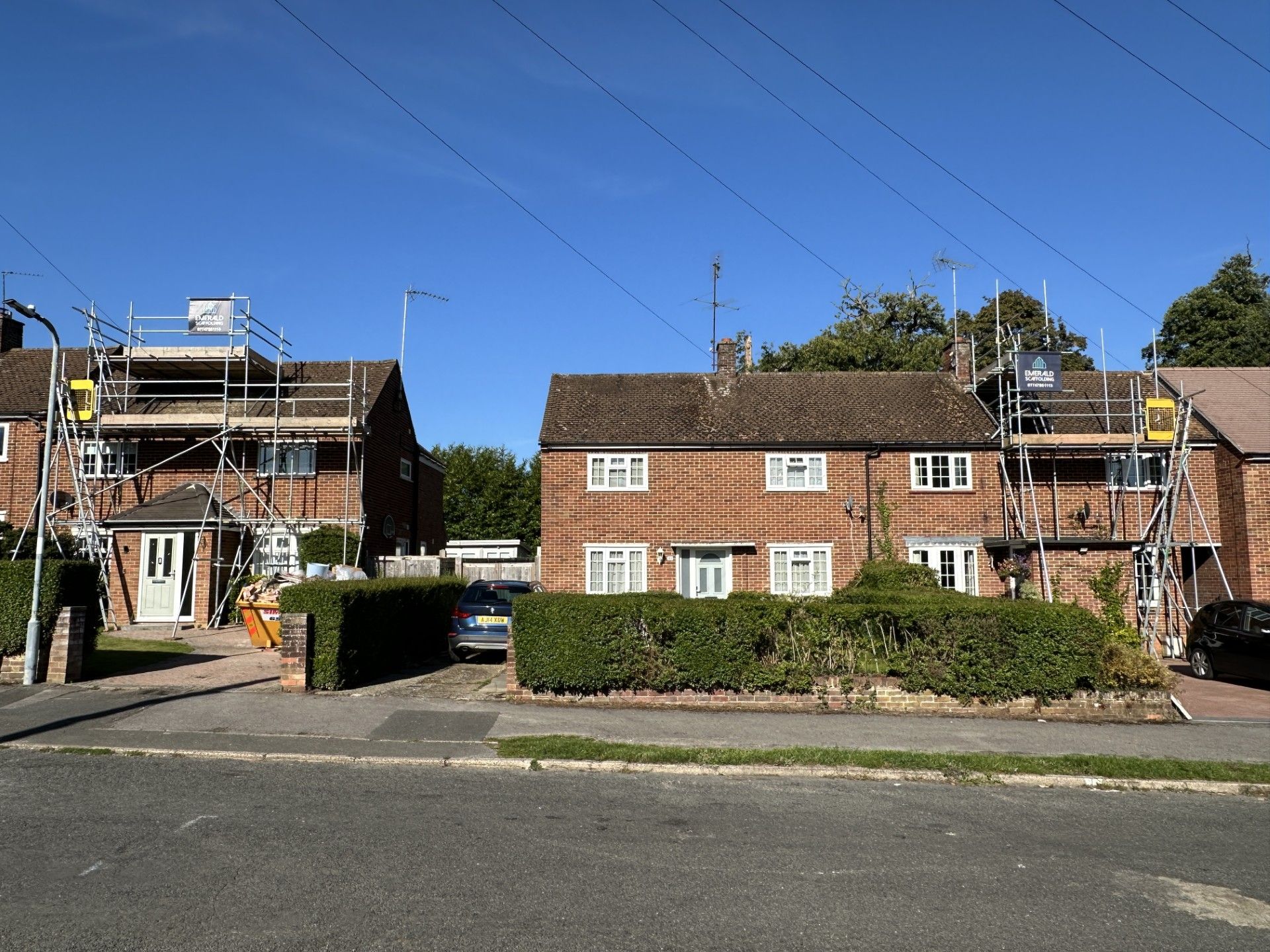 Brick houses with scaffolding, hedges, and parked cars under a blue sky.