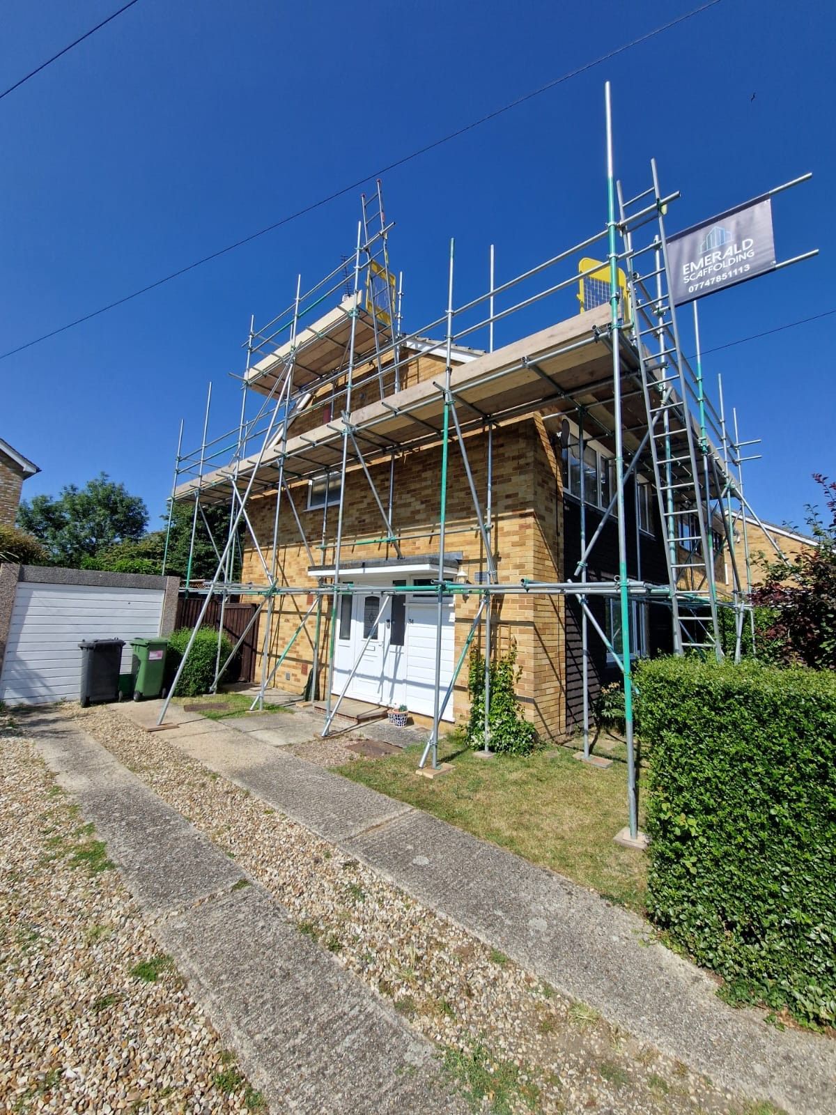 House with scaffolding, driveway, and green lawn under a blue sky on a sunny day.
