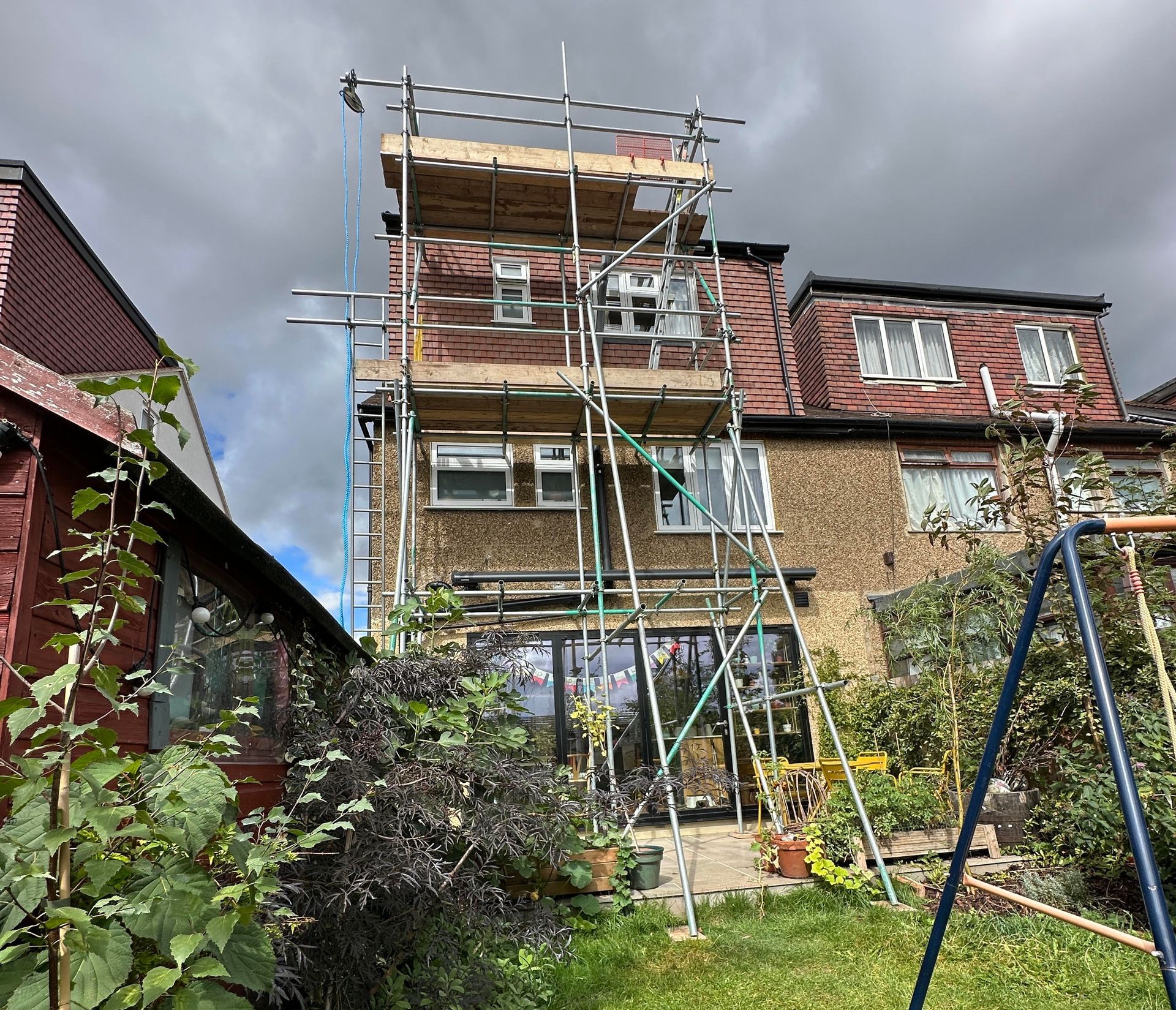 Scaffolding on a brick house with visible windows. Backyard with greenery and a swing set. Cloudy sky.
