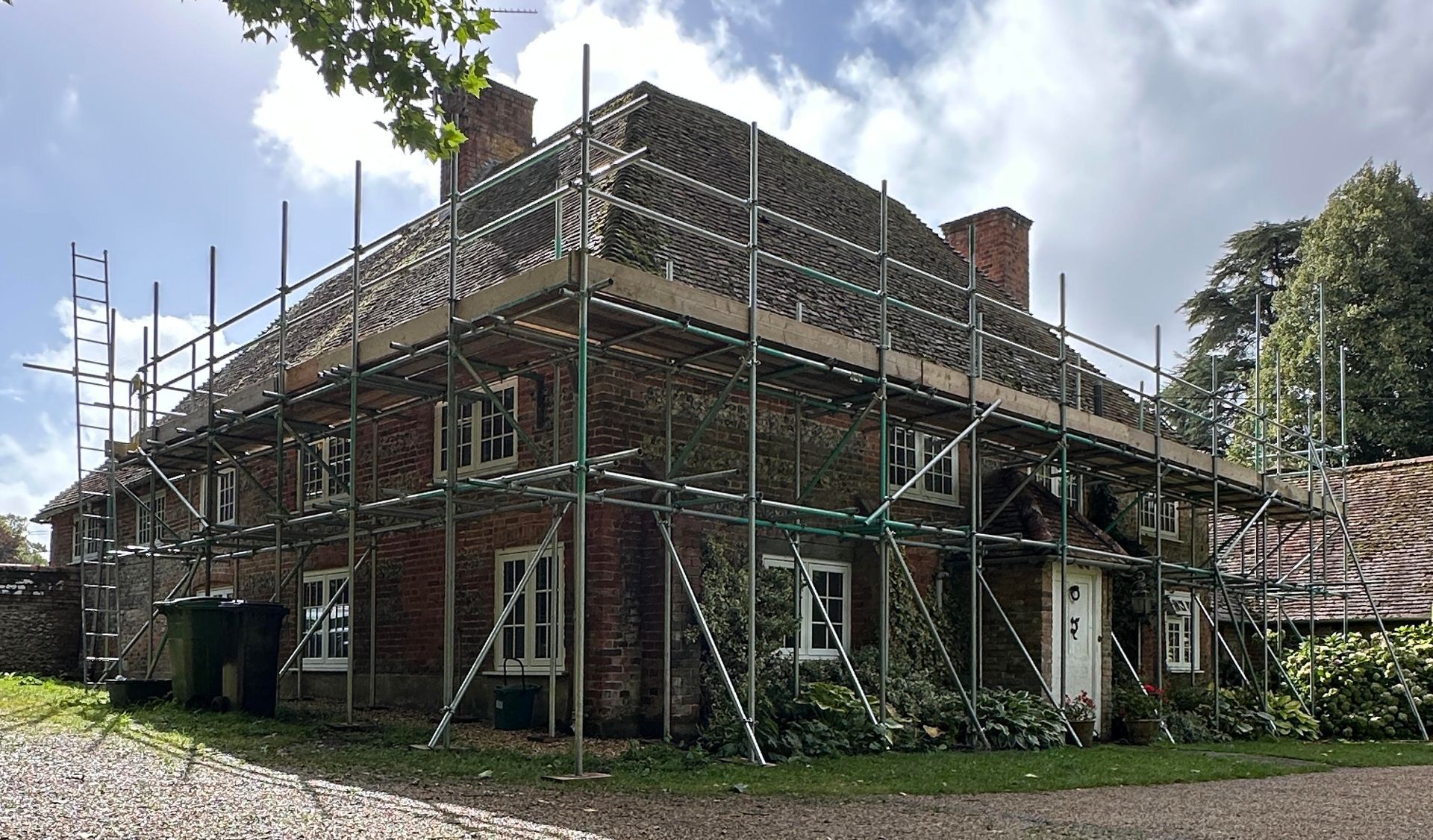Brick house with scaffolding, under construction. Green foliage, trees in background, cloudy sky.